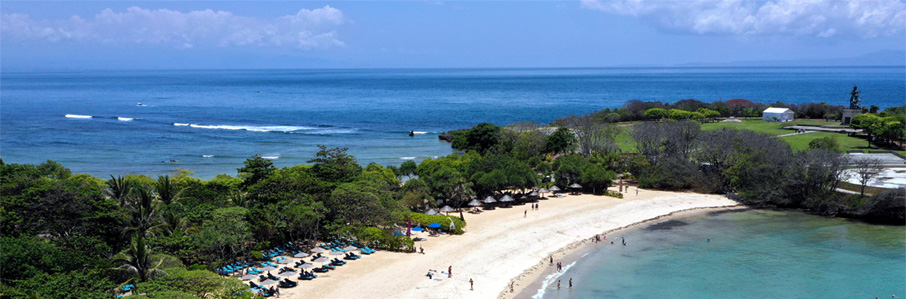 An aerial landscape view of Nusa Dua beach Bali with blue sky above the blue ocean with a strip of beach running from the bottom left of the photo to towards the top right. The white sandy beach has a beackdrop of green trees with beach umbrellas giving shade to blue sunbeds. In the foreground to the bottom right waves gently lap onto the beach.